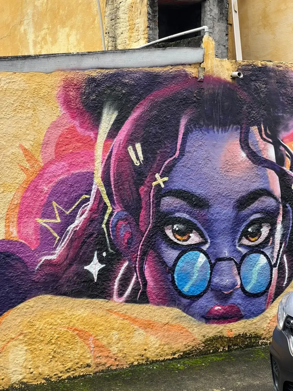Detalhe de mural com rosto feminino em roxo, rosa e azul em uma parede da Vila Flávia.