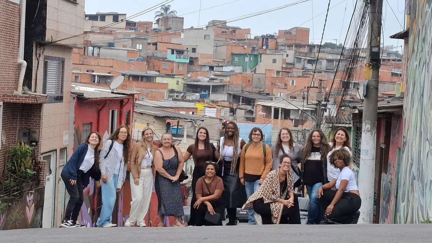 Mulheres reunidas numa rua em declive, com casas da Vila Flávia ao fundo.